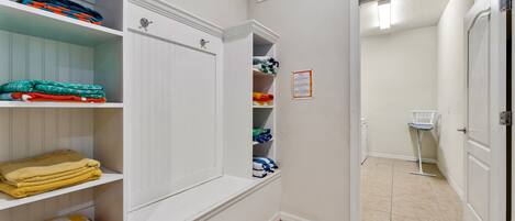 A hallway with white walls, a tiled floor, and a door leading to a bathroom. Shelves are stocked with folded towels and blankets.