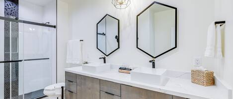 A modern bathroom with a glass shower enclosure, a white toilet, two sinks, and a large mirror.