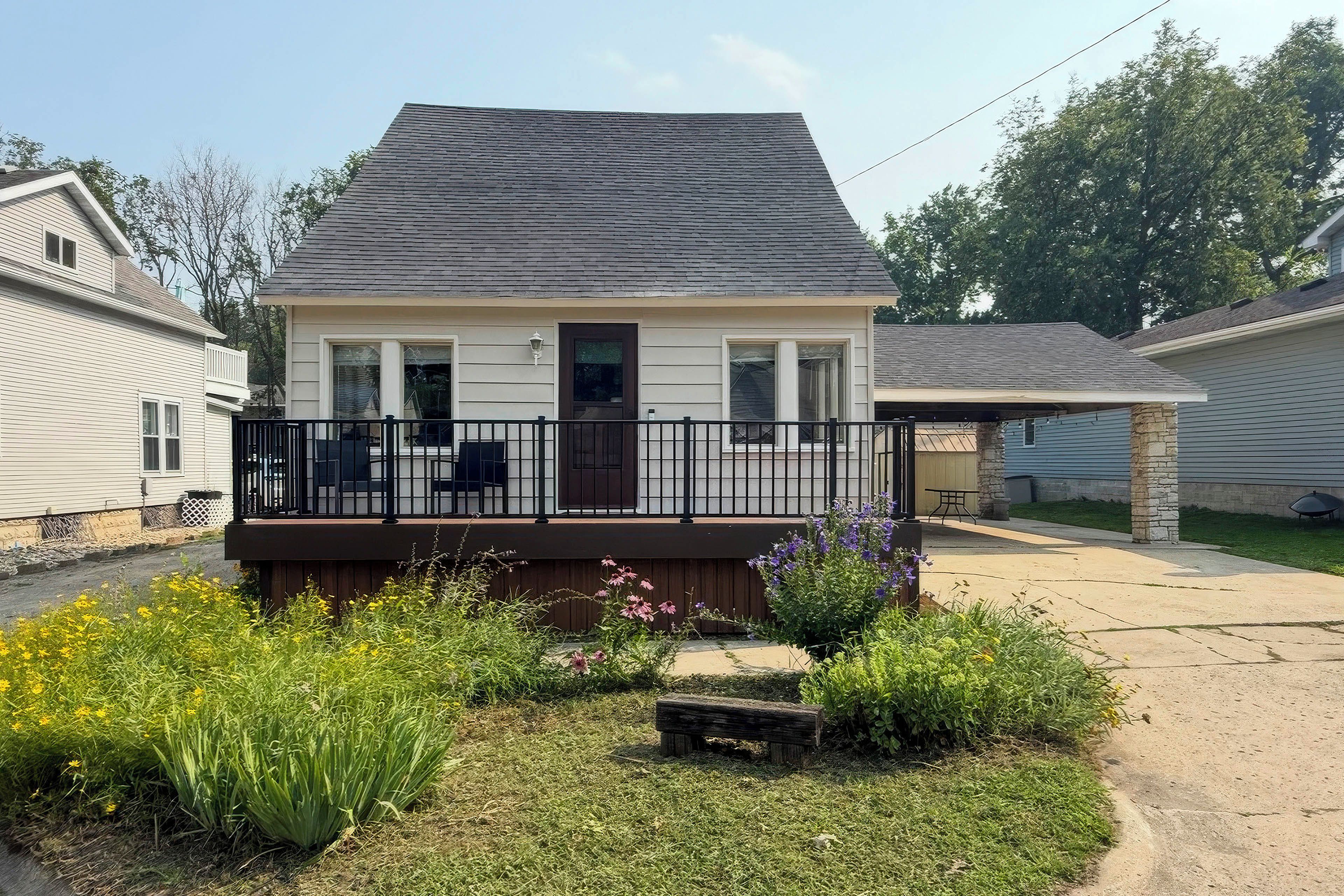 A single-story house with a dark roof, a front porch with a railing, and a small garden with various plants and flowers.
