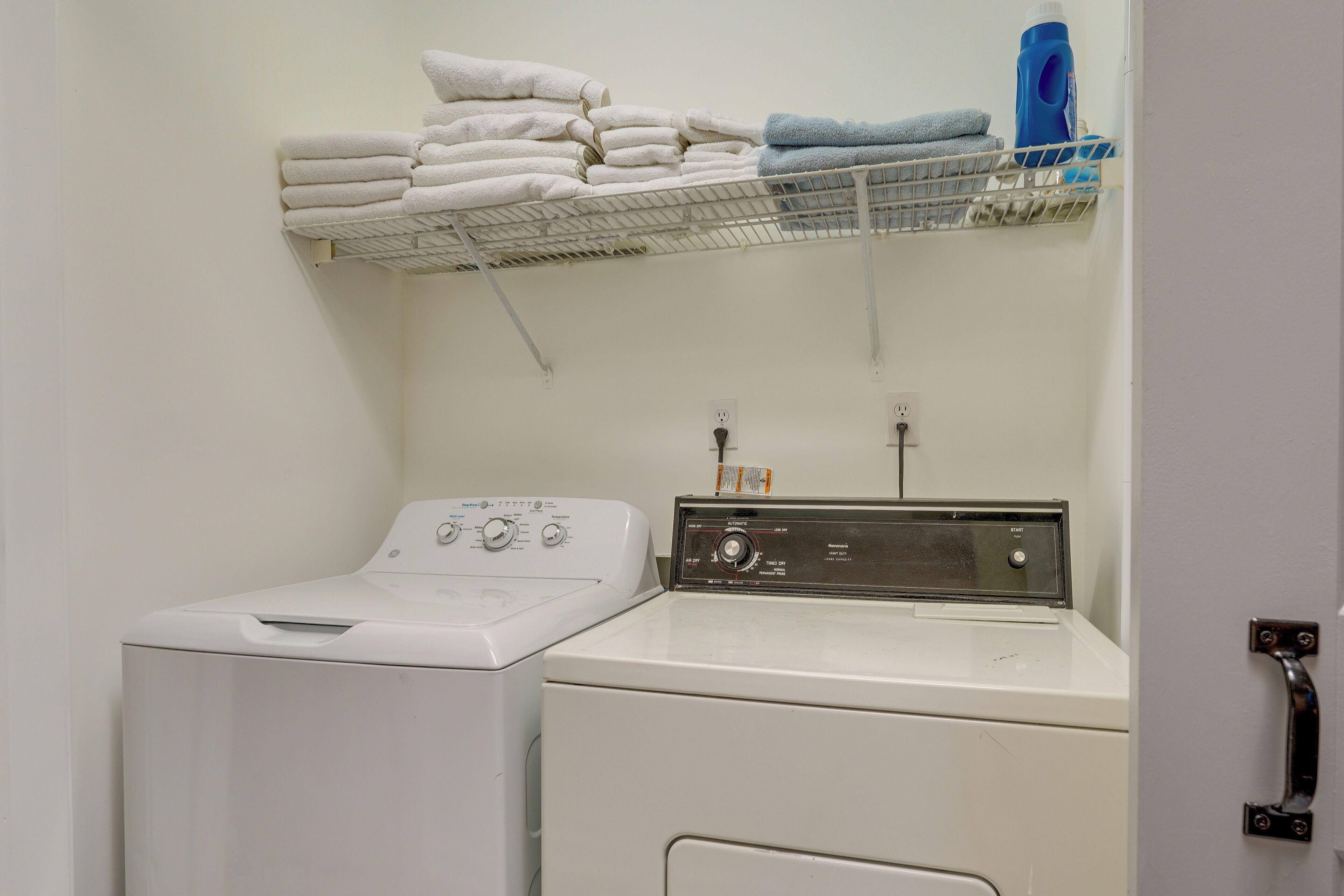 Un cuarto de lavandería con lavadora y secadora de carga frontal, un estante con toallas dobladas y una botella de detergente azul.