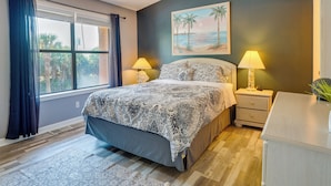 A bedroom with a bed, two bedside tables with lamps, a nightstand, a window with curtains, and a painting of a beach scene. - Walk to Beach: PCB Condo w/ Oceanfront Pool Access (Panama City Beach)