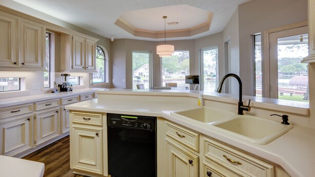 Una cocina con muebles blancos, lavavajillas, pileta doble y ventanas amplias.