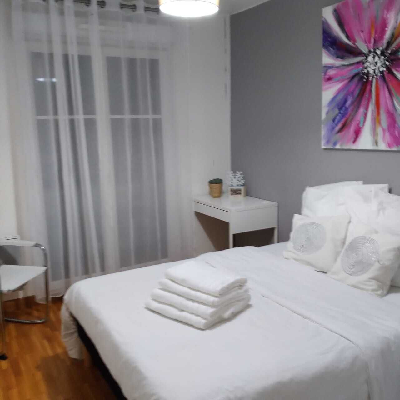 A neatly made bed with white linens, a bedside table with a small plant, and a large abstract painting on the wall.