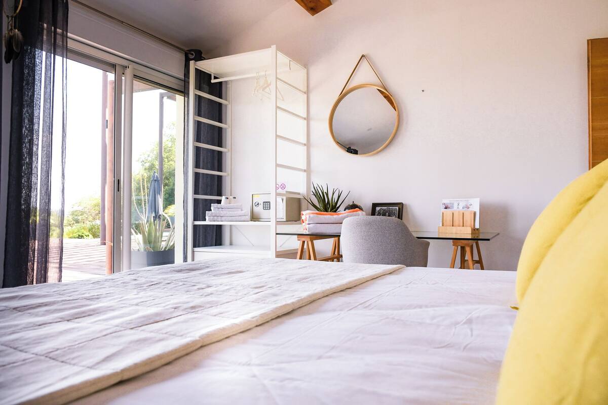 Een moderne slaapkamer met een groot bed, een bureau met stoel, een hangspiegel en een schuifpui naar een balkon.