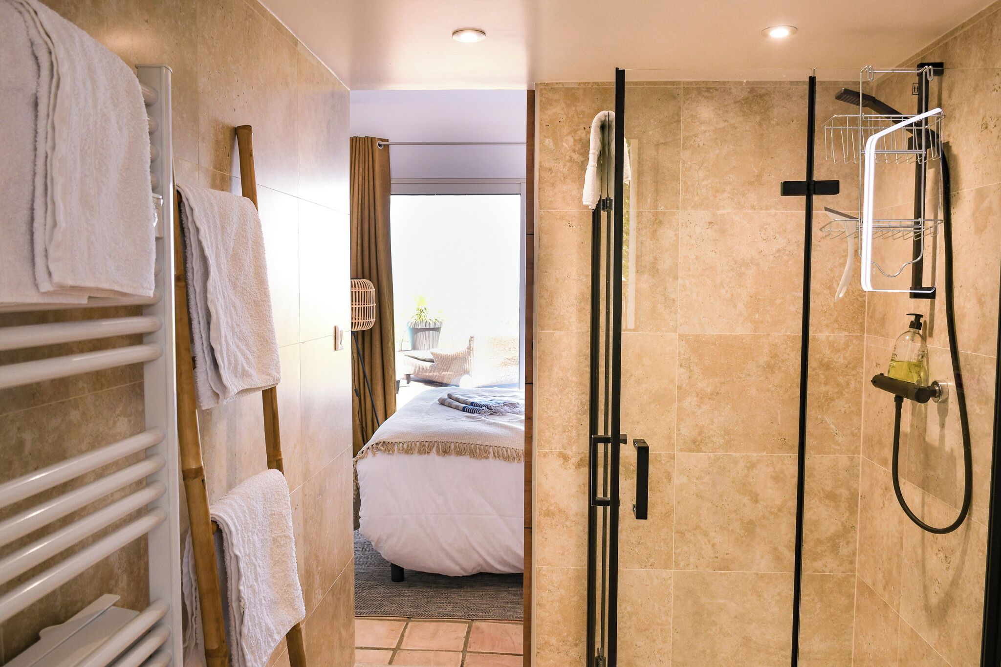A hotel bathroom with a shower, a towel rack, and a bed visible through an open door.
