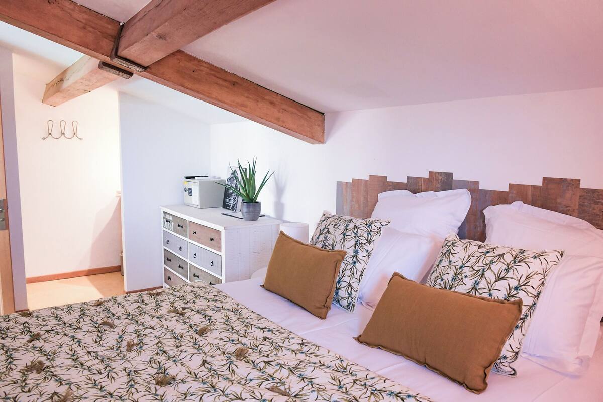Een slaapkamer met een bed, witte muren, een houten hoofdbord, een dressoir, een plant en een raam.