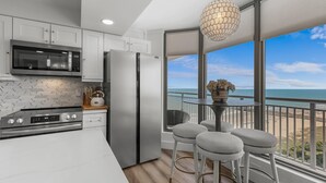 A modern kitchen with stainless steel appliances, a white countertop, and a large window overlooking a beach. - 1606 Brighton 3 Bedroom Condo (Myrtle Beach)