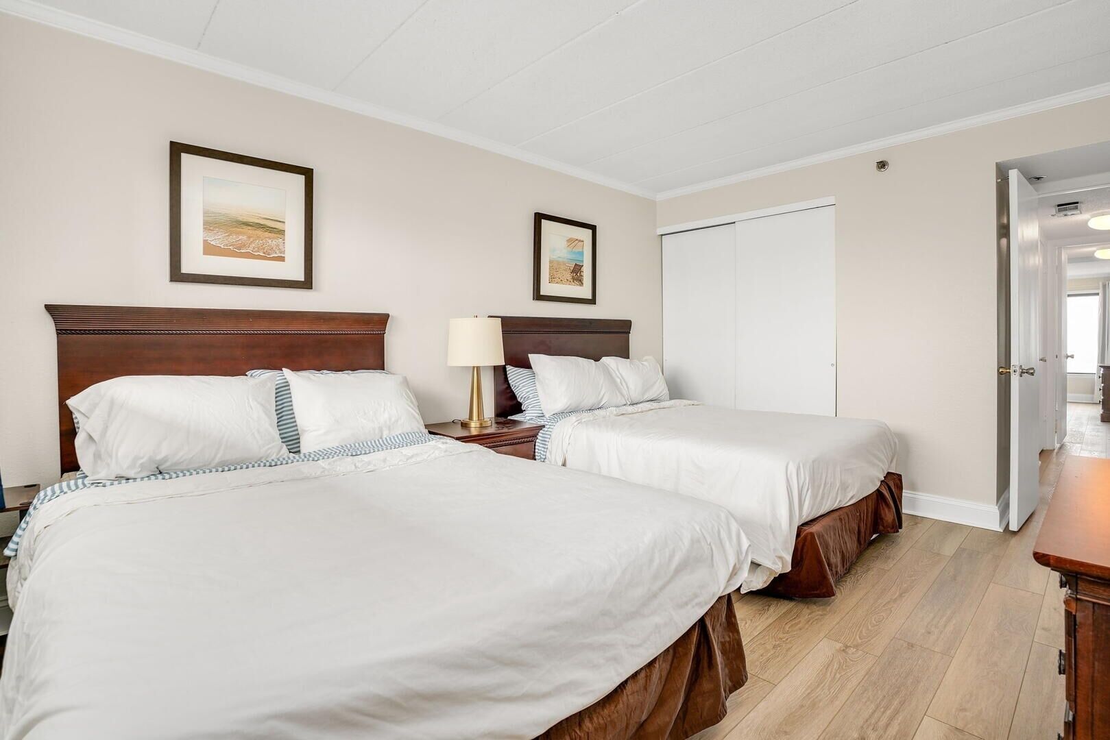Une chambre d’hôtel avec deux lits, une tête de lit en bois, une table de chevet avec une lampe et une porte donnant sur une autre pièce.