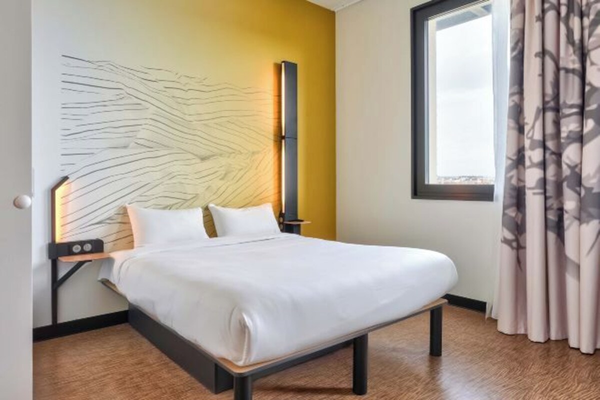 Een moderne hotelkamer met een bed, een nachtkastje, een kunstwerk aan de muur en een raam met gordijnen.
