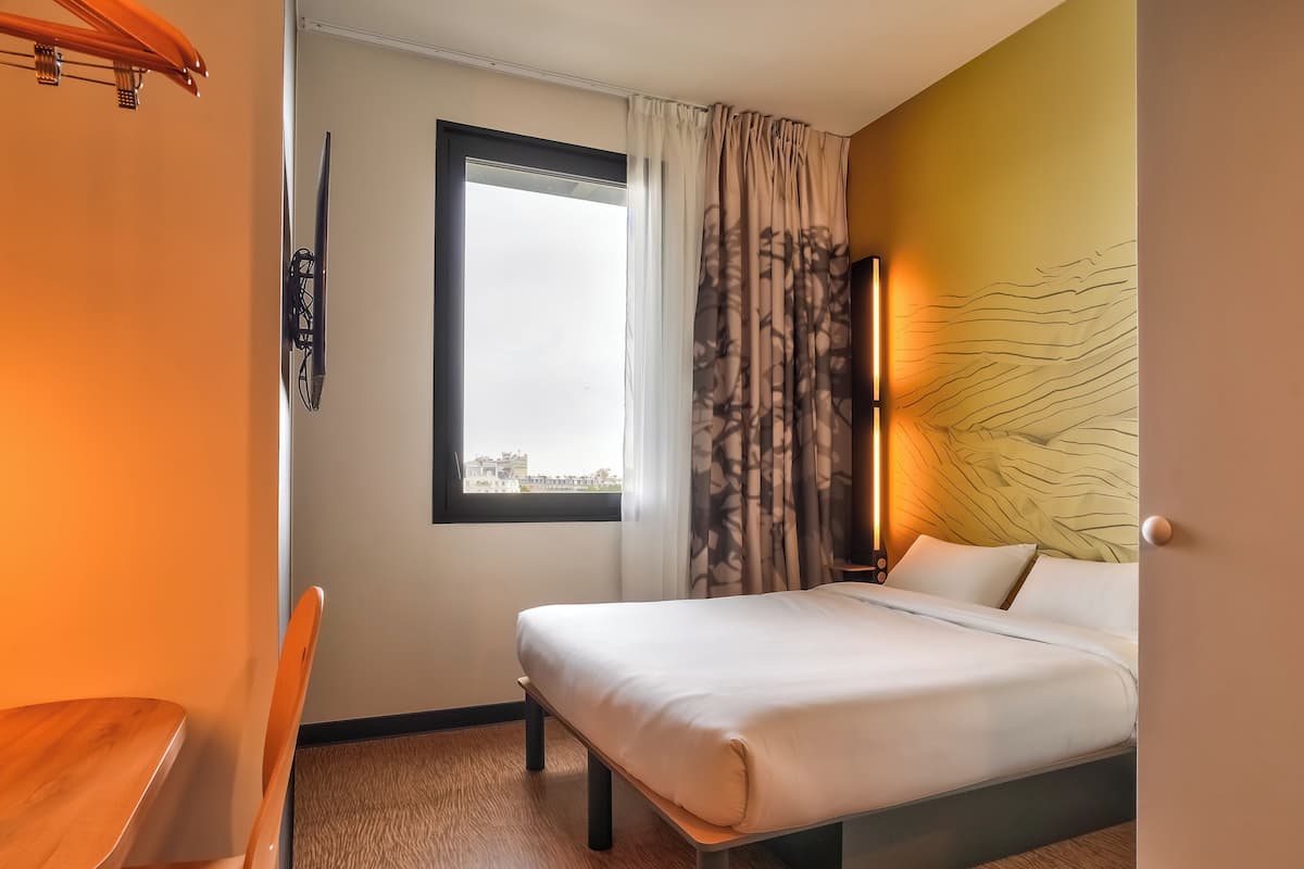 Hotellihuone, jossa on sänky, ikkuna verhoilla, seinään kiinnitetty valaisin ja näkymä ulkona oleviin rakennuksiin.