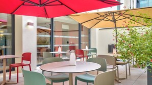 Een terras met tafels en stoelen, rode en beige parasols, en uitzicht op het interieur van het gebouw.