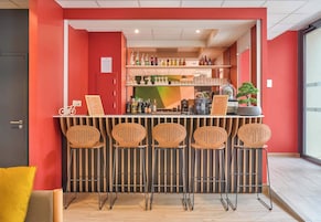 Uma área de bar com parede vermelha, várias prateleiras cheias de garrafas e cinco banquetas.