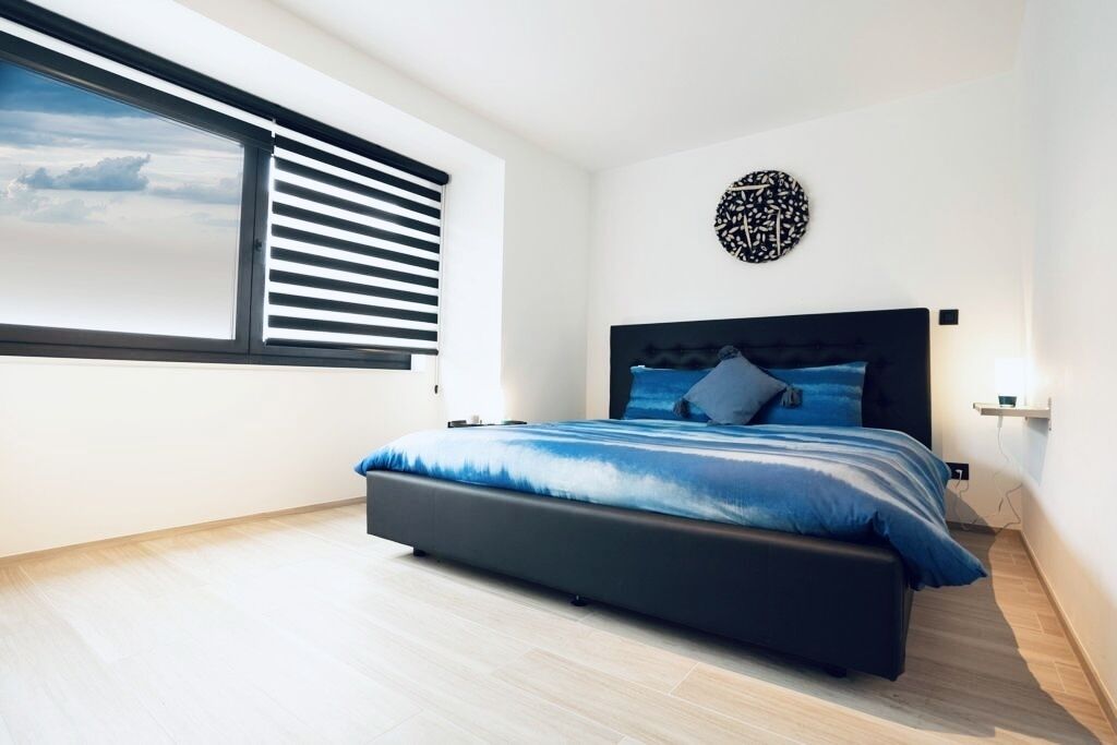 Une chambre moderne avec un grand lit, une fenêtre avec des stores, une horloge murale et un interrupteur.