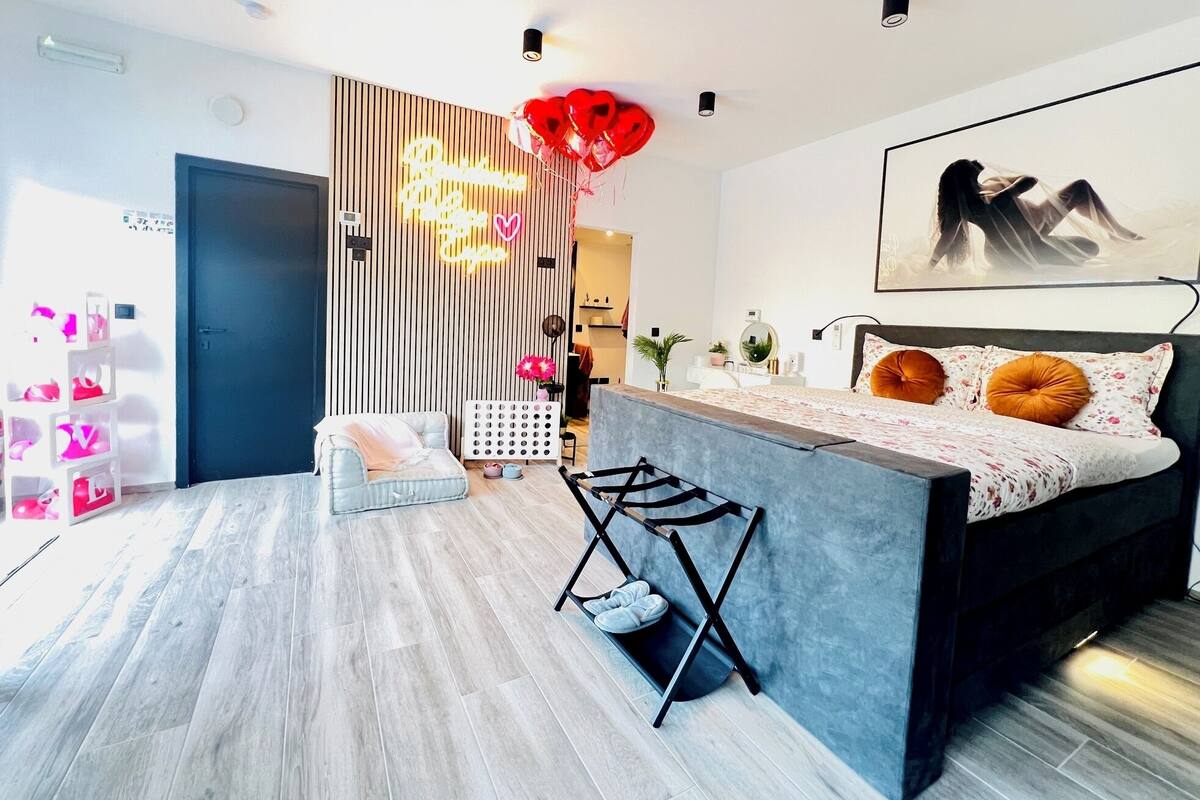 Une chambre moderne avec un lit, un bureau et une chaise. Un néon est accroché au mur, et il y a aussi un tableau.