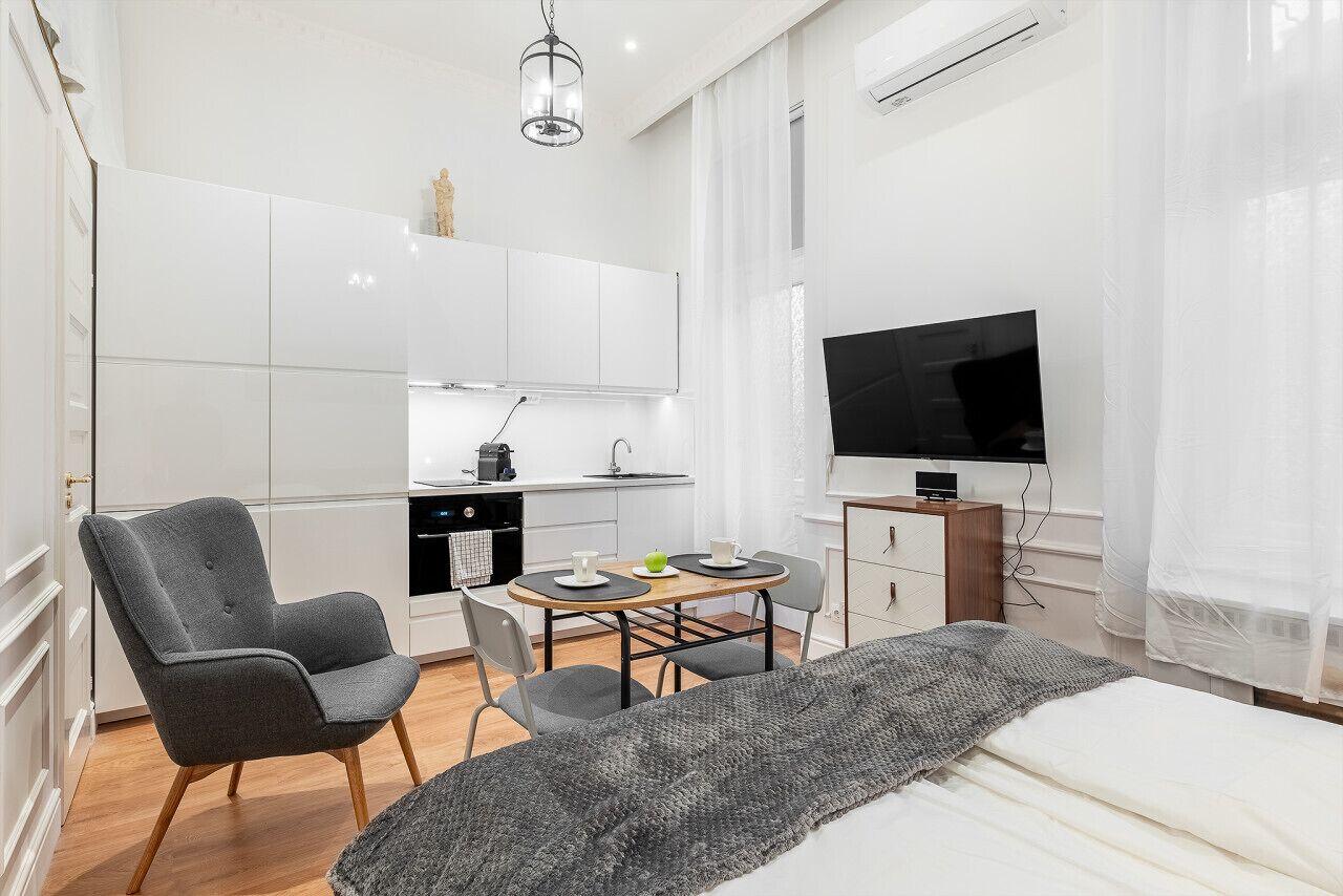 A modern bedroom with a bed, a grey armchair, a small dining table with chairs, a TV, and a kitchenette in the background.