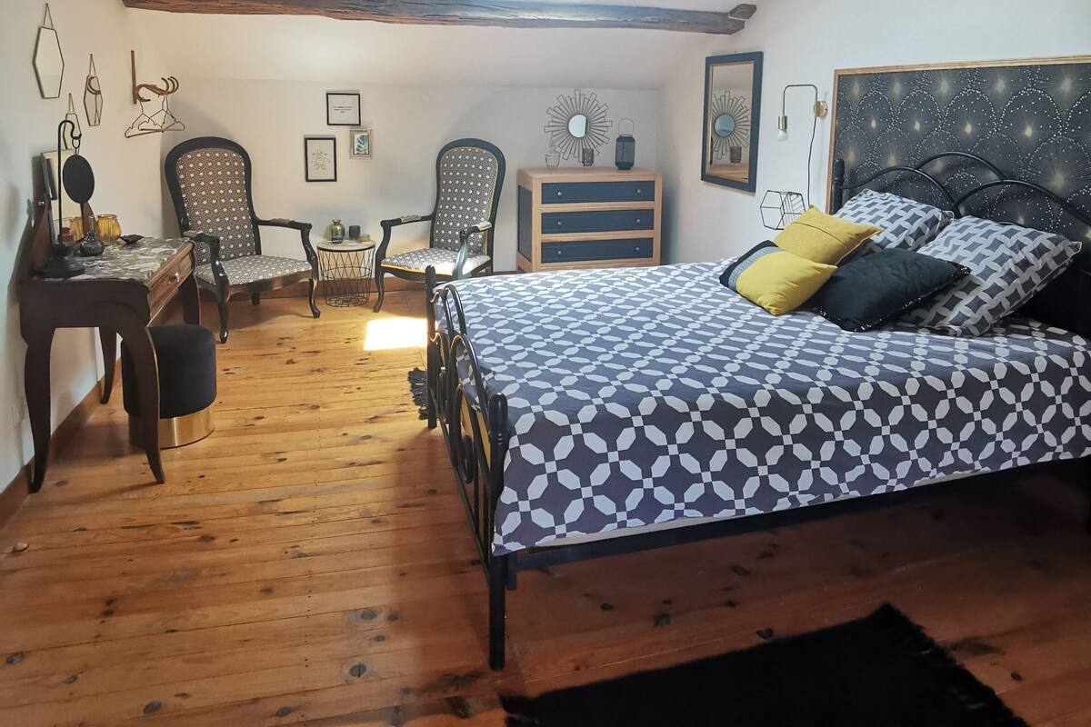 A cozy bedroom with a sloped ceiling, a bed with patterned bedding, a desk with a chair, and a small table with decorative items.
