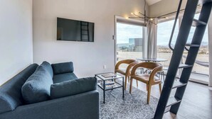 A modern living room with a grey sofa, a glass coffee table, and a TV mounted on the wall. There is a window with a view of the desert and a ladder leaning against the wall. - Stardust Luxury Cottage with loft & a great view (Terlingua)
