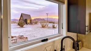 A window view of a rustic cabin with a metal roof, surrounded by dry vegetation and mountains in the background. - Stardust Luxury Cottage with loft & a great view (Terlingua)