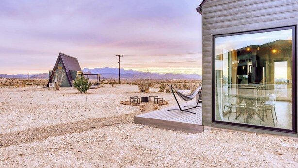 A modern building with a large glass window, a hammock, and a small table with chairs in a desert setting. - Stardust Luxury Cottage with loft & a great view (Terlingua)