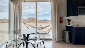A room with a large window, a dining table with chairs, a hammock, and a kitchenette. - Stardust Luxury Cottage with loft & a great view (Terlingua)
