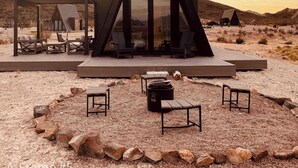 Un edificio moderno de forma triangular con fachada de vidrio, rodeado por una terraza con asientos al aire libre y un pozo de fuego.