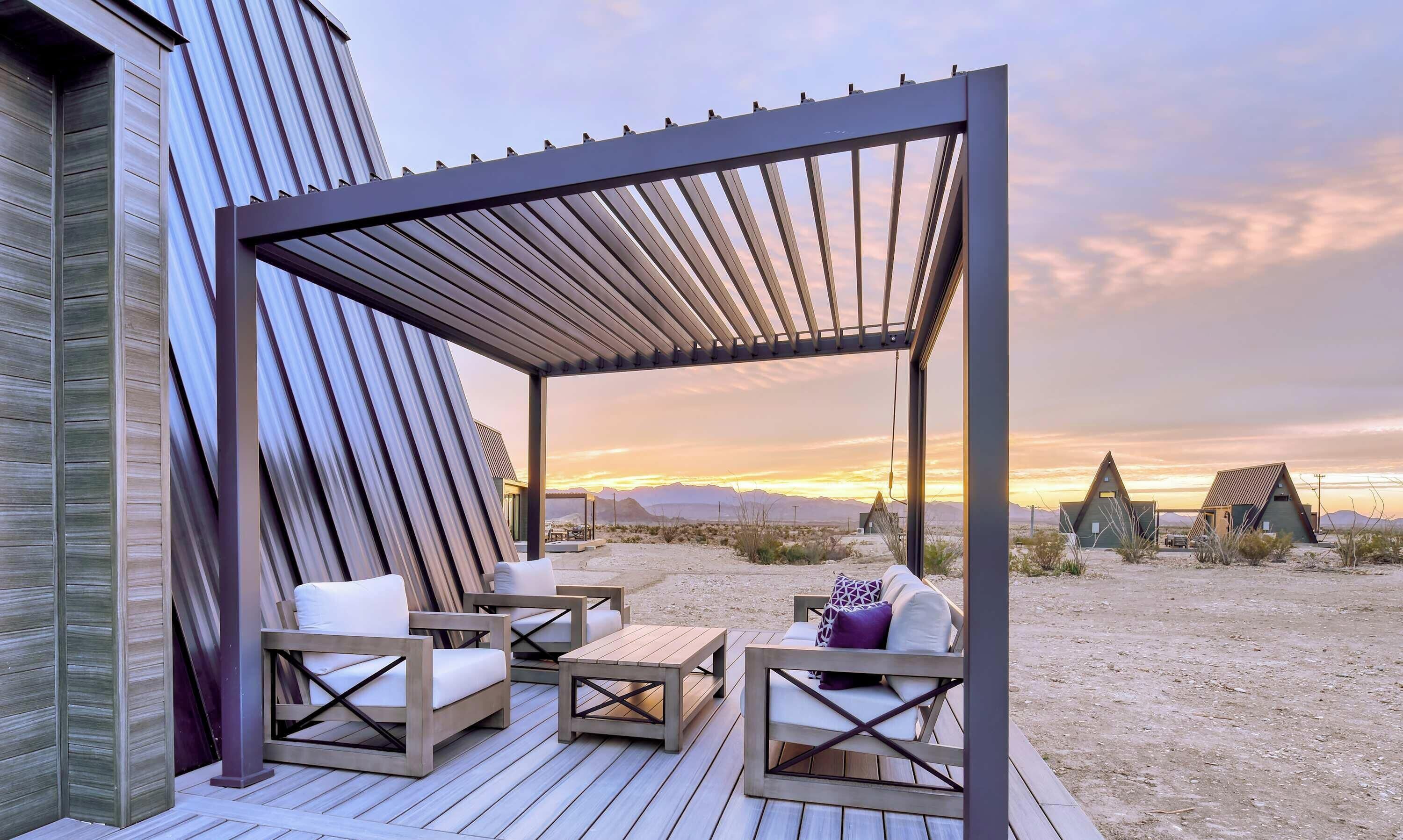 Una terraza de madera con mobiliario exterior, una pérgola y vistas a edificios en el desierto al atardecer.