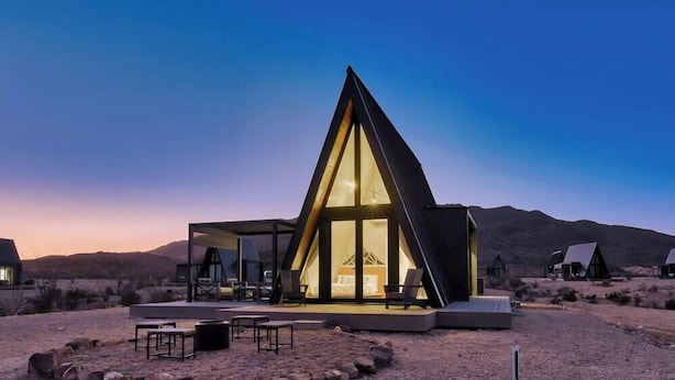 A modern, triangular-shaped building with a glass facade, outdoor seating area, and a view of the surrounding desert landscape. - Stardust Big Bend Luxury A-Frame#10 with fab view (Terlingua)
