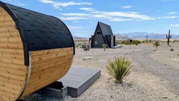A wooden barrel-shaped structure with a metal roof, a small building with a triangular roof, and a desert landscape with mountains in the background.