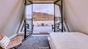 A bedroom with a bed, a chair, and a view of a beach through a glass door. - Stardust Big Bend Luxury A-Frame#8 with a fab view (Terlingua)
