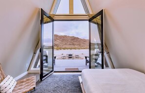A bedroom with a bed, a chair, and a view of a beach through a glass door. - Stardust Big Bend Luxury A-Frame#8 with a fab view (Terlingua)