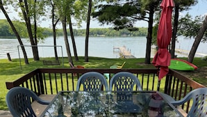A deck with a glass table and chairs, overlooking a lake with a dock and chairs on the grass.
