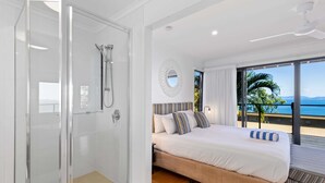 A modern bedroom with a large bed, a glass-enclosed shower, and a balcony with a view of the ocean.
