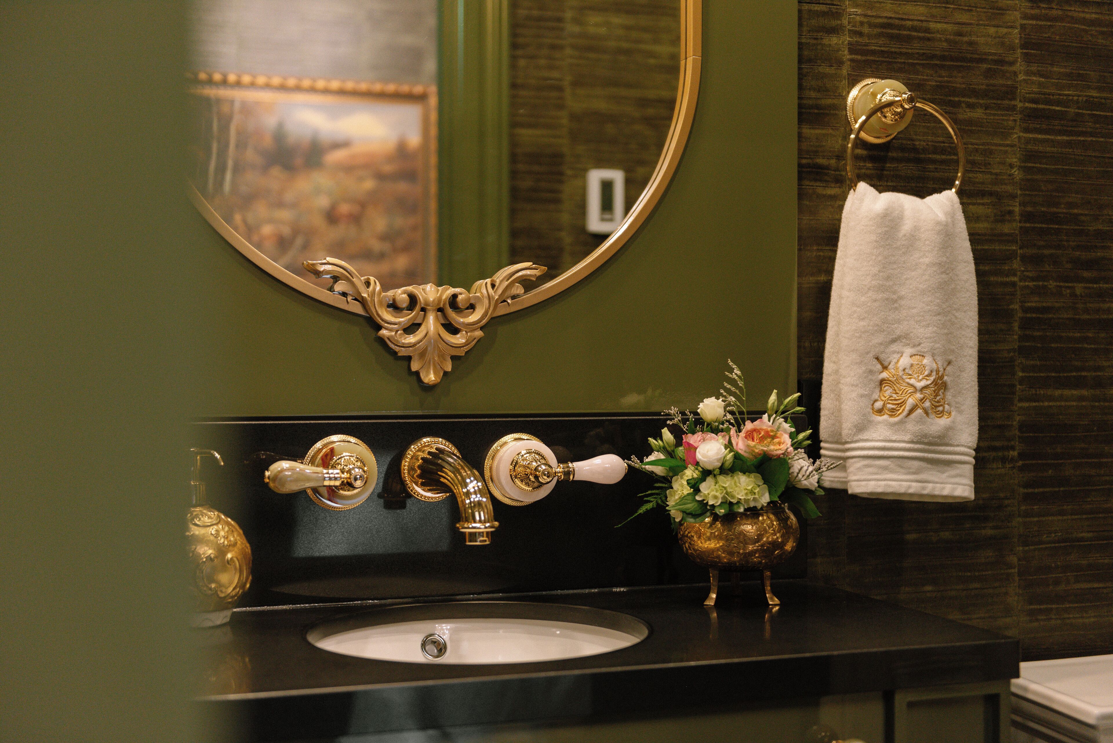Ein Badezimmer mit einem spiegelnenden Wandspiegel mit goldener Rahmen, einem schwarzen Waschbecken und einem Wasserhahn in Goldfarbe.