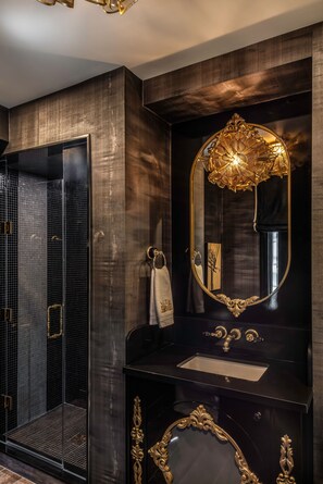 A modern bathroom with a large mirror, dark wood paneling, a glass shower enclosure, and a gold-framed vanity. - The Scotsman Hotel (Niagara-on-the-Lake)