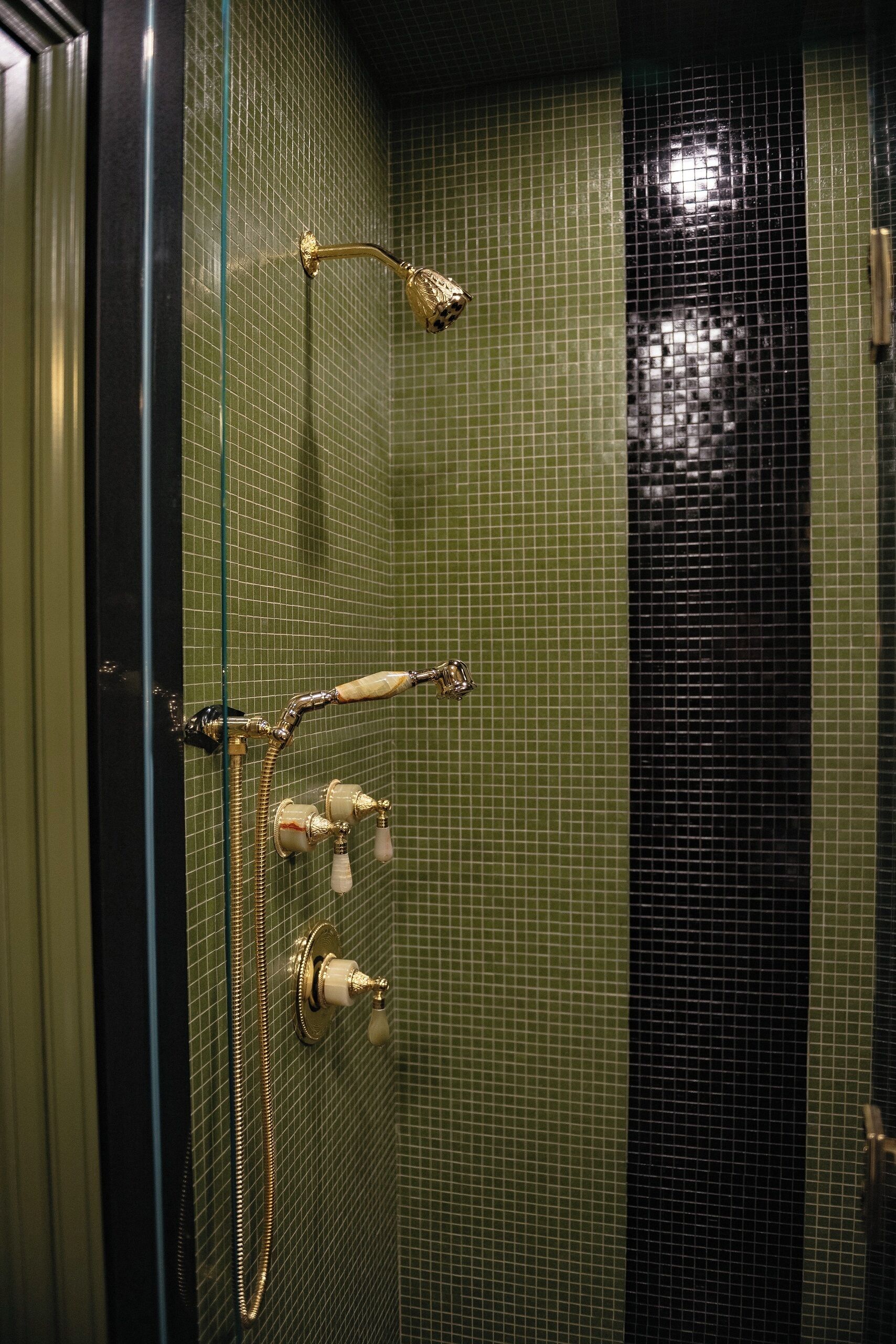 Ett badrum med en vägg täckt av gr öna mosaikplattor, ett gyllene duschmunstycke och en handdukshängare.