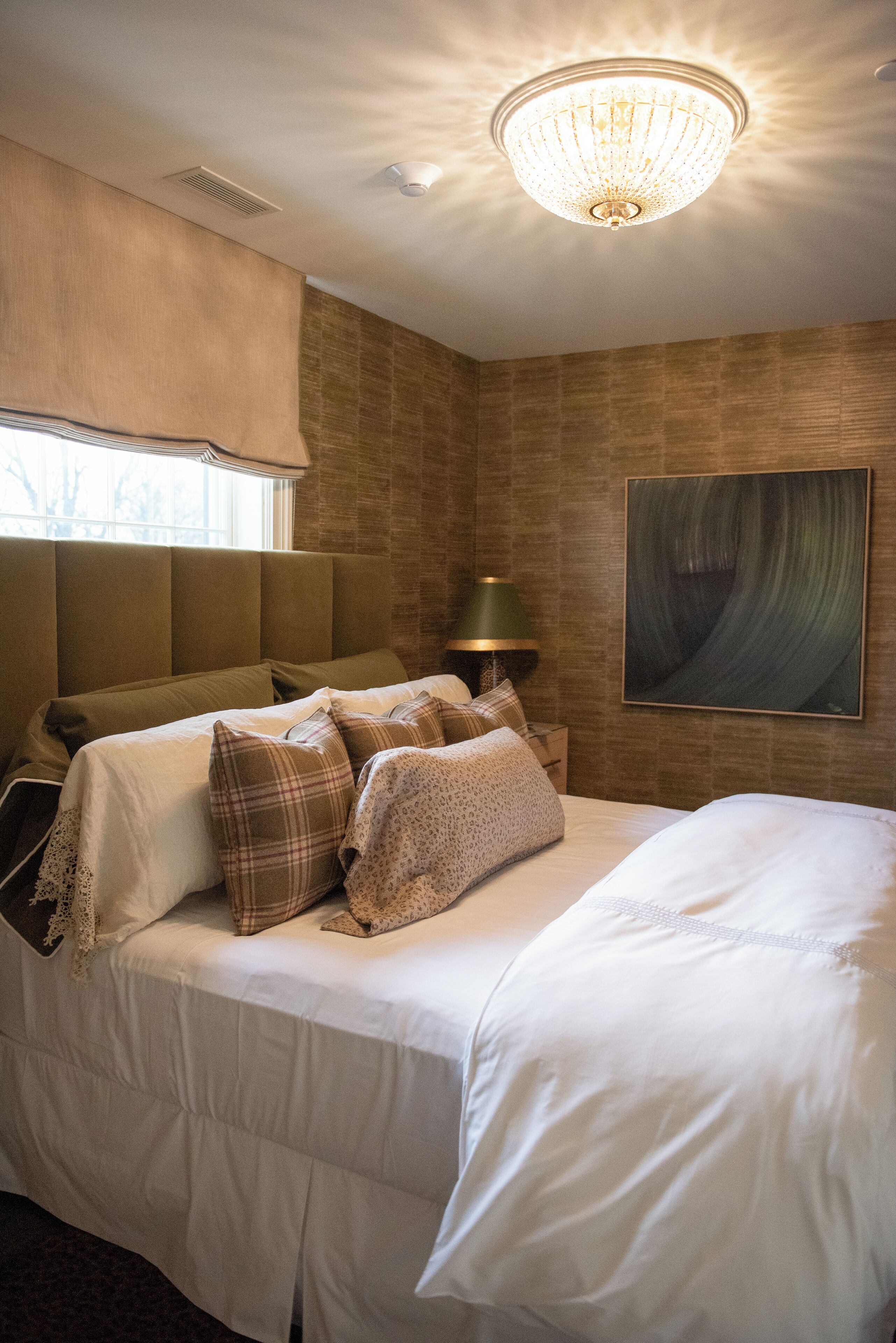 Een net opgemaakt bed met hoofdbord, een bedlampje en een schilderij aan de muur.