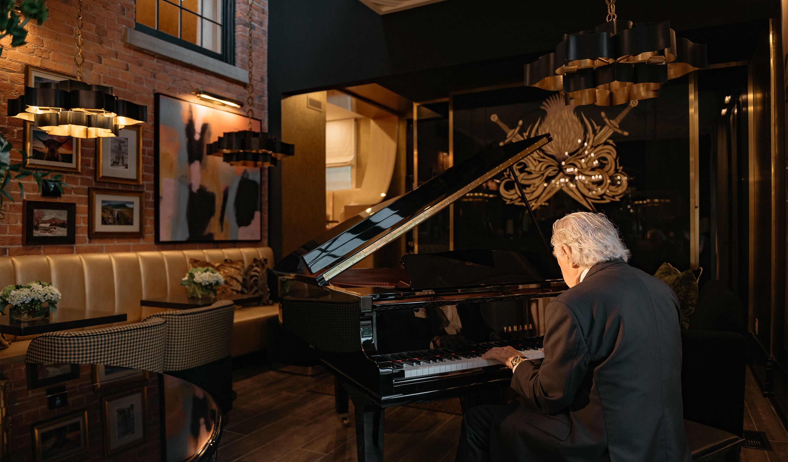 A man playing a grand piano in a warmly lit room with a brick wall, framed artwork, and a chandelier.