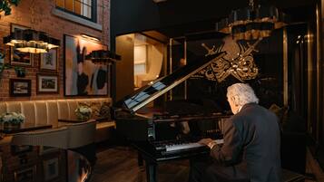A man playing a grand piano in a warmly lit room with a brick wall, framed artwork, and a chandelier.