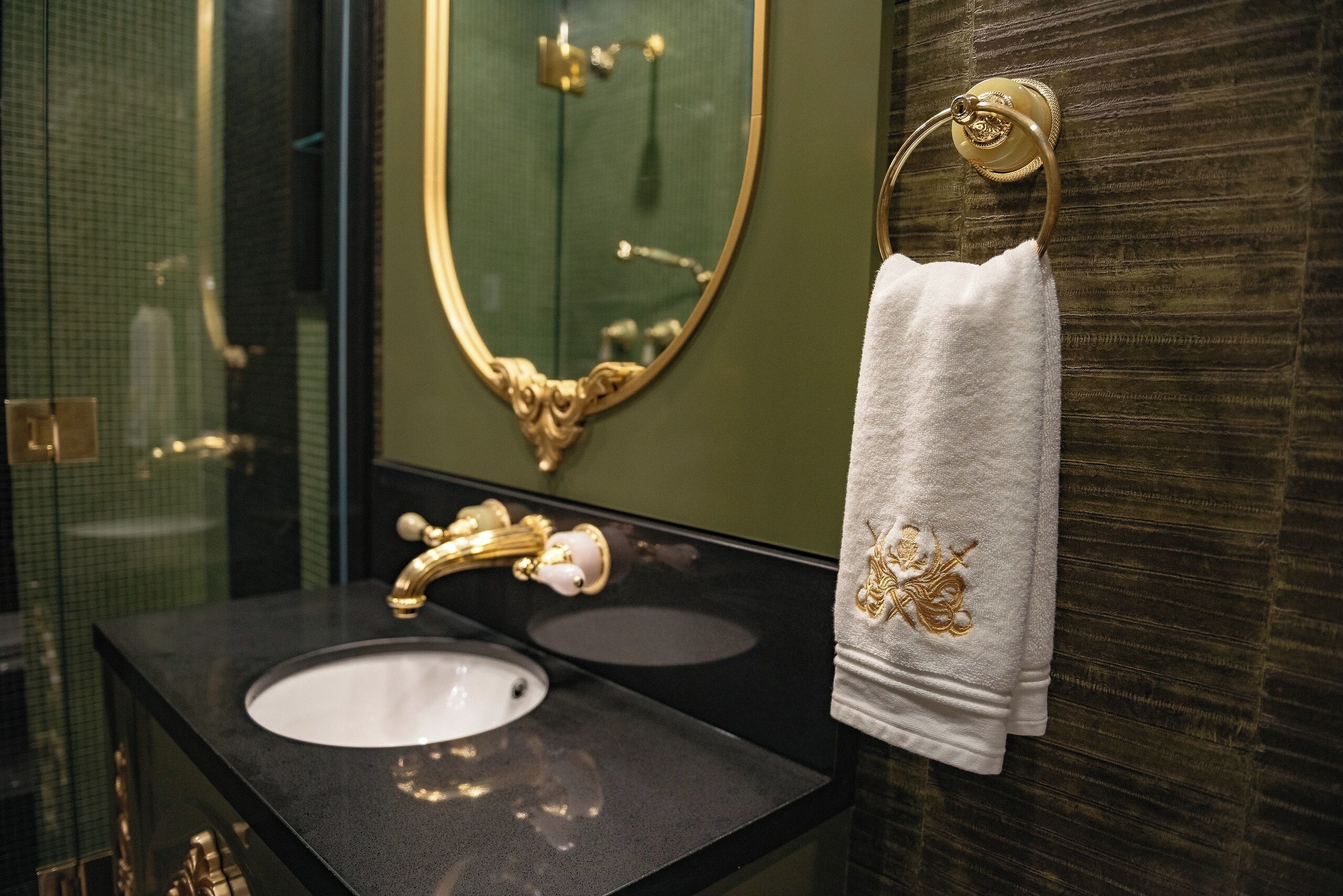 A bathroom with a black countertop, a gold faucet, a white sink, and a towel with gold embroidery.