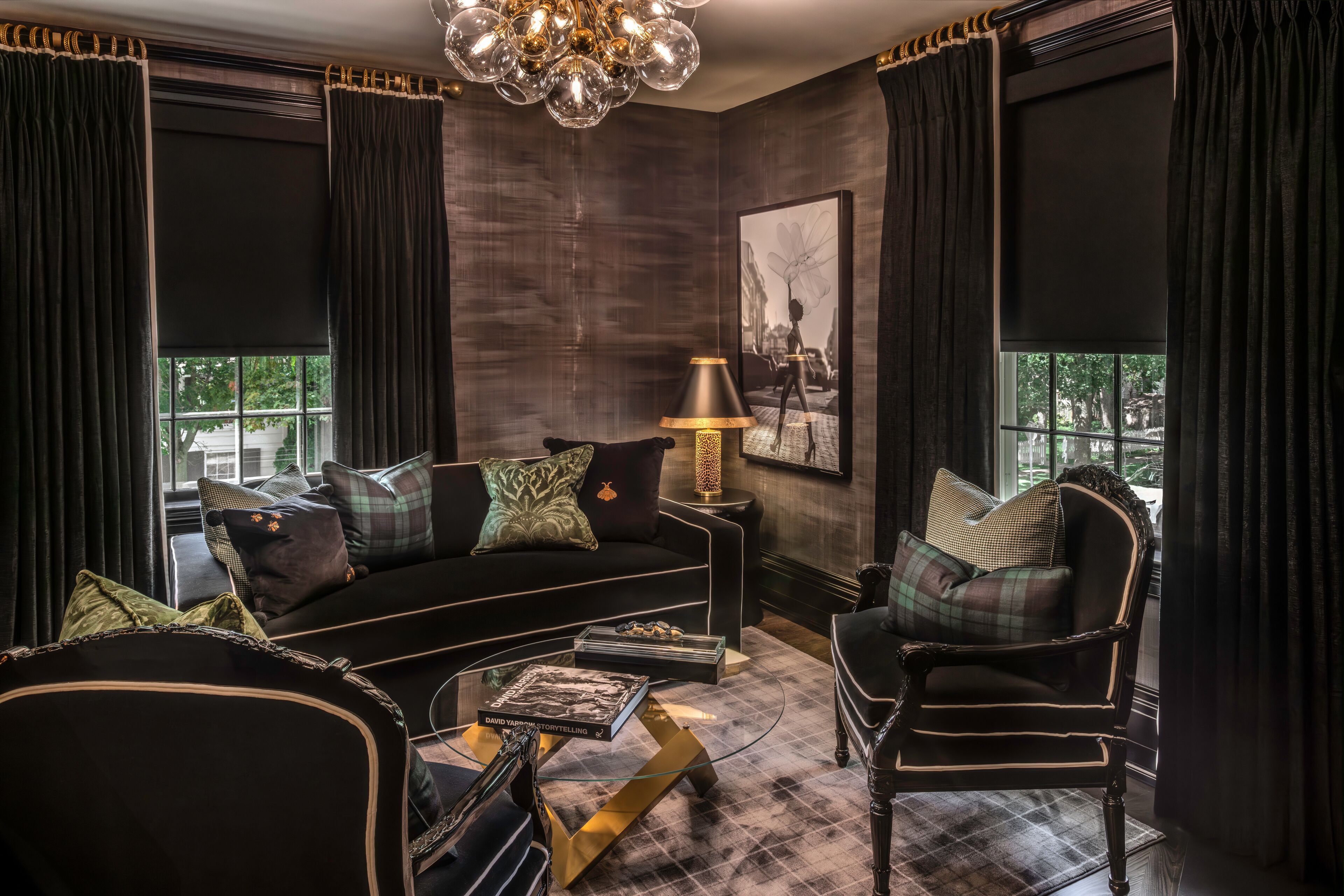 Un salón con decoración oscura, que incluye un sofá negro, una mesa de centro de cristal y un sillón oscuro. Una lámpara y un cuadro enmarcado están colgados en la pared.