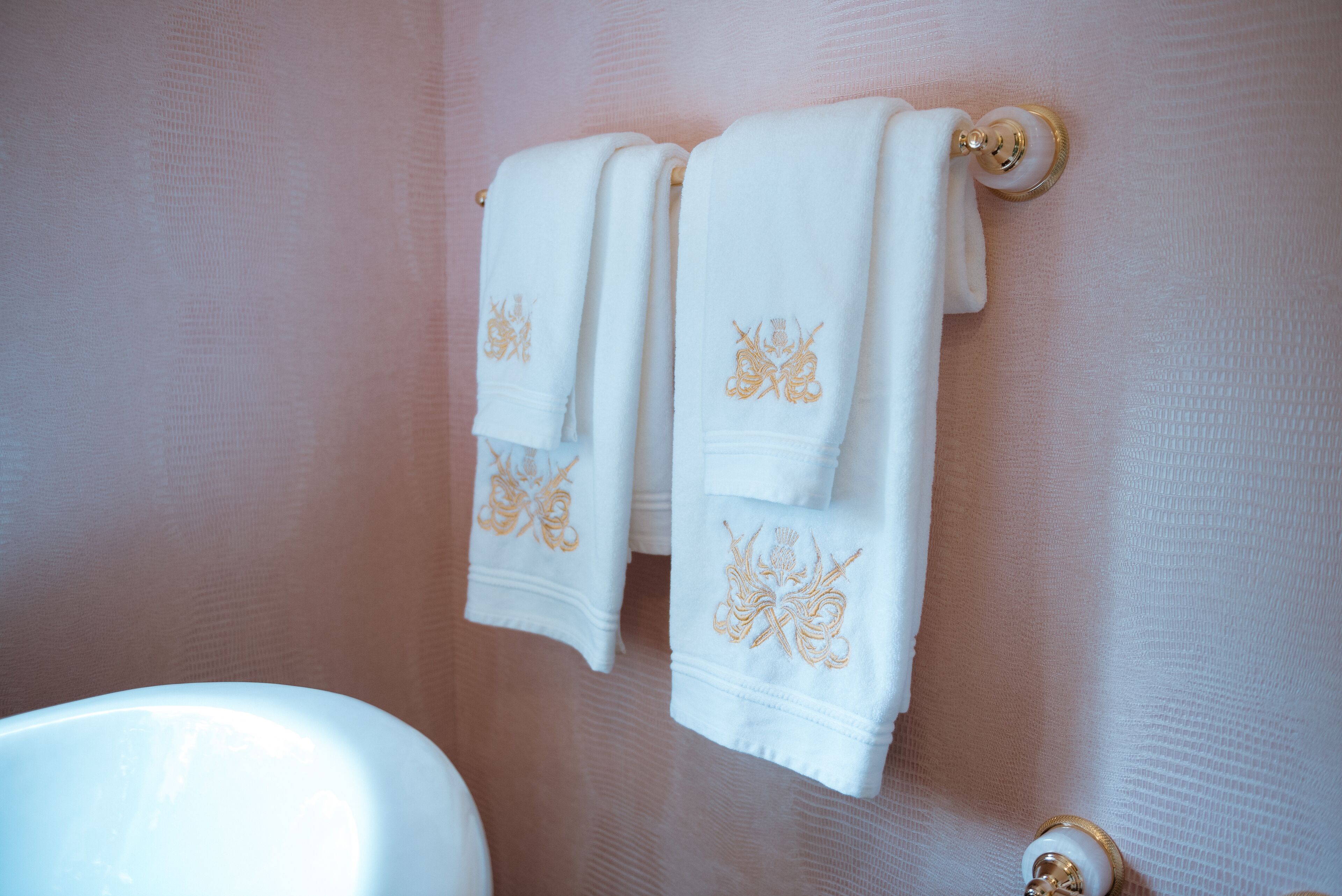 Uma casa de banho com uma banheira branca, duas toalhas brancas com bordados e um suporte de toalha dourado.