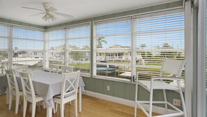 Un espace repas comprenant une table et des chaises, de grandes fenêtres équipées de stores et un ventilateur de plafond.