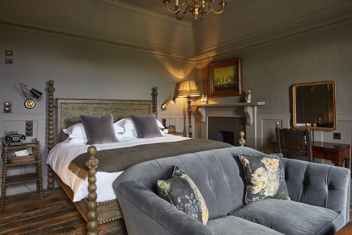 A bedroom with a large bed, a grey sofa, a chandelier, a fireplace, and a mirror.