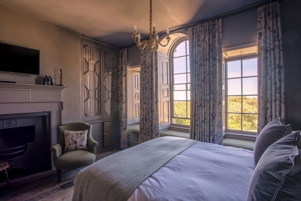 A bedroom with a large bed, a fireplace, a television, and a view of the outdoors through a window.