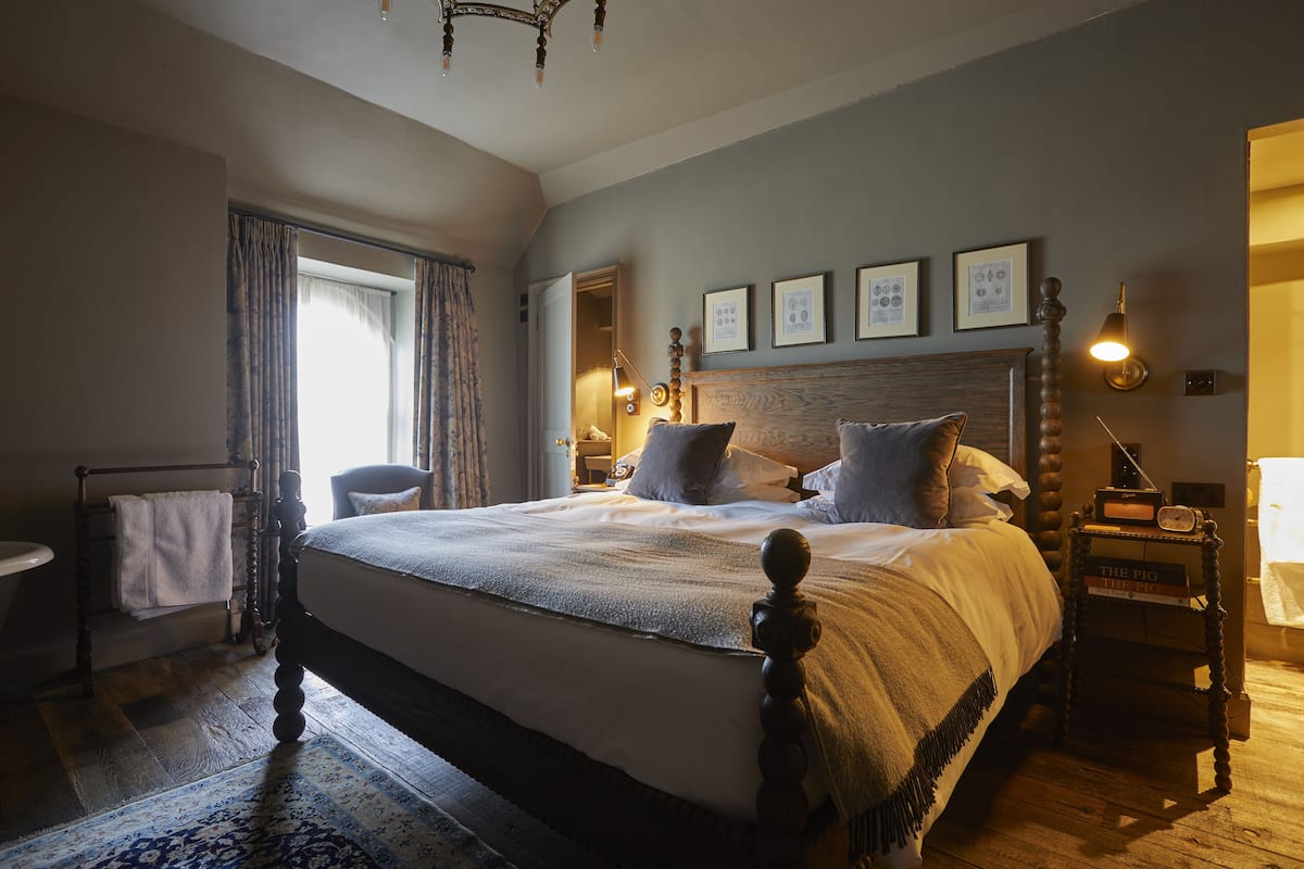 A bedroom with a large bed, a bedside table with a lamp, a chair, and a window with curtains.