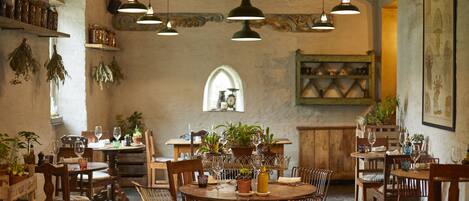 A rustic dining area with wooden tables and chairs, hanging pendant lights, and potted plants.