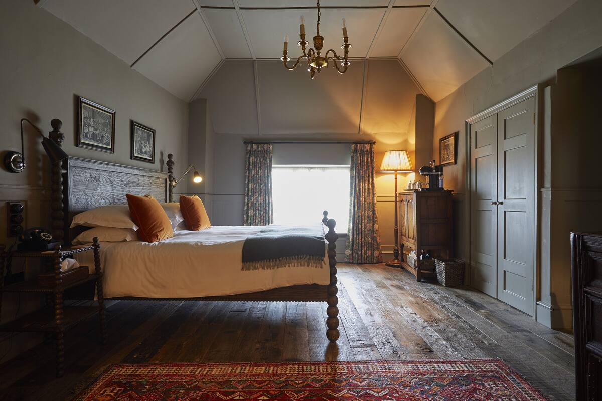 A bedroom with a large bed, wooden floor, a chandelier, a nightstand, and a window with curtains.