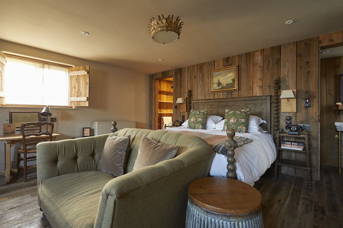 A cozy bedroom with a wooden headboard, a green armchair, a round wooden stool, and a desk with a chair.