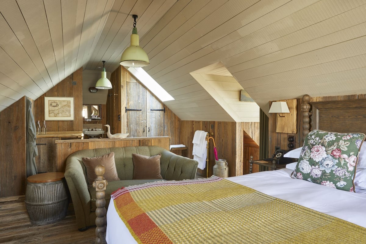 A rustic attic bedroom with a wooden ceiling, a bed with a floral pillow, a green sofa, and a small table with a lamp.