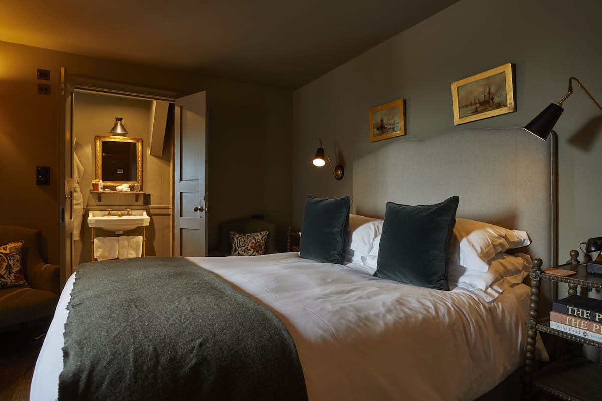 A neatly made bed with a white comforter and dark throw, a bedside table with books, a lamp, and a small shelf with a mirror and towel.